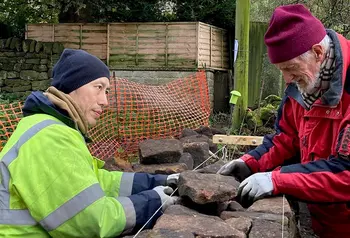 A person demonstrates dry stone wall construction techniques to two people in an outdoor setting