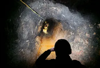 A rock walled mineshaft illuminated by torchlight stretched into the distance, in the foreground the silhouette of a person wearing a miners helmet and looking along the tunnel