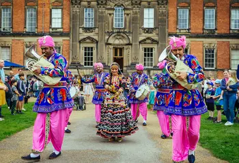 An Asian brass band, with musicians and dancers dressed in colourful clothes, walking down a path in front of a stately home