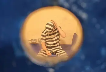 A photo taken through a keyhole of a prisoner in a cell, wearing striped black and white clothing.