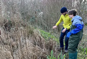 Two people look at an overgrown pond, one person is wearing waders