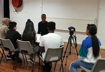 A group of young people sit a table interviewing another young person, with another person to the side operating a camerawith 