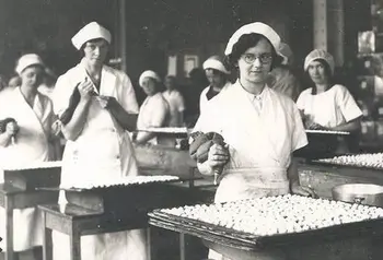 Women wearing uniforms, stood at tables in a factory, making biscuits.