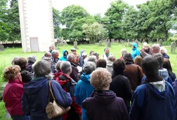 A group of people wearing outdoor clothes take part in a churchyard heritage walk at St Matthew's Churchyard