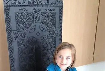 Young child sitting on a high backed wooden chair depicting an intricate carving of a cross. 