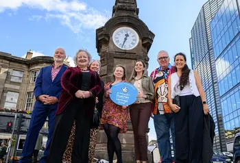 A project group of seven people pose for a photograph in front of the Hear of Midlothian Memorial Clock. Karen Mailley stands at the centre of the group, holding a Heritage Fund branded roundel.