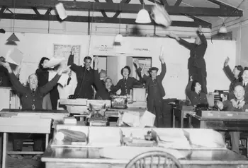 A black and white photograph showing RAF staff celebrating in an office, throwing their arms and papers in the air.