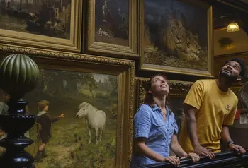 Two people stand on a staircase looking up, with paintings covering the wall behind them
