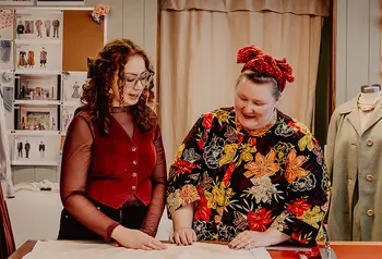Two people are in a costume room and are looking down at a pattern on a cutting table. In the background are images of costumes, costumes on mannequins and rolls of fabric stacked on shelves