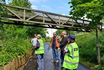 A group of people standing in a lane above them is a wooden bridge, the lane is enclosed by greenery covered embankments.