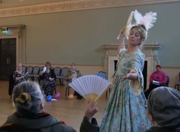 A person wearing an 18th-century style green floral gown and feathered headpiece performs a dance routine to an audience inside a historic hall