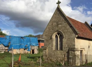 A stone chapel in the foreground, a derelict timber-framed building covered in plastic sheeting in the background