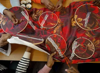 A top down view of several people's hands sewing on a large piece of patterned fabric covered in embroidery hoops