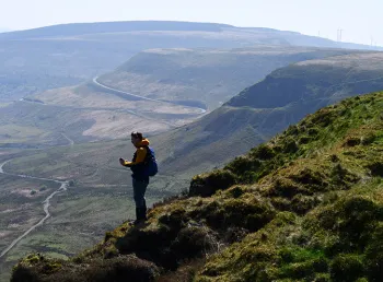 A person wearing a yellow coat with a blue backpack stands on a hillside surrounded by rolling Welsh hills