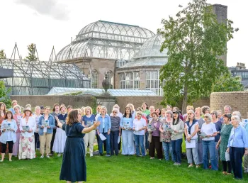 a choir sing in a garden in front of a Victorian glass and ironwork Palm House