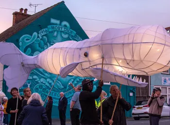 A large whale-shaped lantern being paraded in front of a mural of Moby Dick
