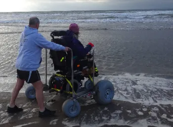 Man pushing person using large wheelchair next to river