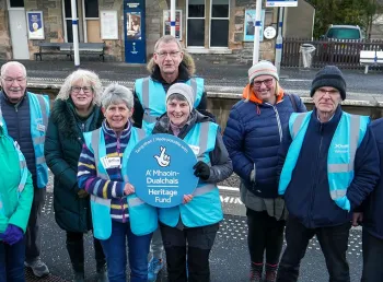 A group of people wearing cold weather gear stand on the platform of a rural train station holding a sign 