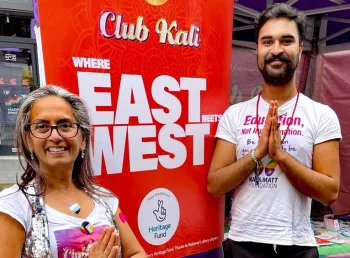 Two south asian people stand next to a banner with text: Club Kali Where East Meets West made possible with Heritage Fund