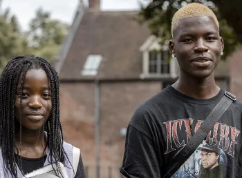 Two young black people wearing casual clothes stand outside with a historic brick building in the background