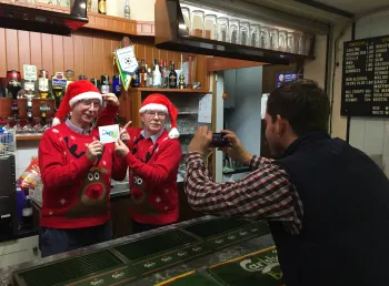 A person taking a photo of two people behind a bar wearing red Christmas hats and jumpers.