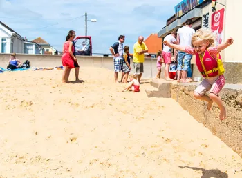 A young girl jumping from a wall onto the sand at Jaywick in Essex. Families are in the background at a cafe.