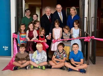 A group of children and adults sit and stand as they pose for a photo while cutting a ribbon.