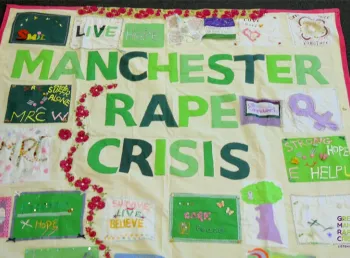 The banner has the words Manchester Rape Crisis in the middle with other phrases and images stitched around it