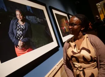 On the left is a framed photograph of a woman on the right there is a woman in a light brown dress looking at the picture