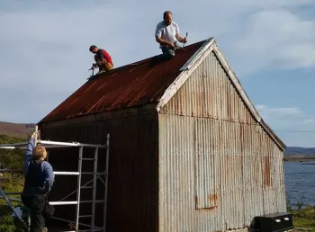 Men working on a small corrugated building with water and mountains in the distance.
