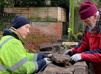A person demonstrates dry stone wall construction techniques to two people in an outdoor setting