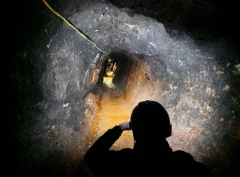 A rock walled mineshaft illuminated by torchlight stretched into the distance, in the foreground the silhouette of a person wearing a miners helmet and looking along the tunnel