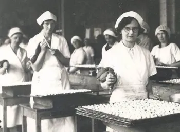 Women wearing uniforms, stood at tables in a factory, making biscuits.