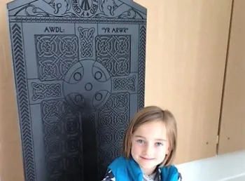 Young child sitting on a high backed wooden chair depicting an intricate carving of a cross. 