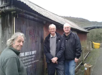 Three people stand outside a large closed shed with a sign saying awaiting restoration
