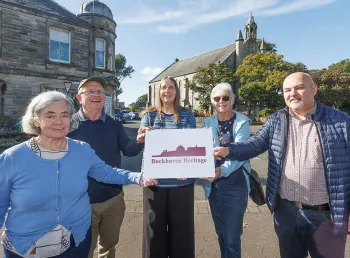 five people holding a Buckhaven Heritage sign. Behind them a stone church can be seen