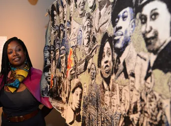 Paula Ogun Hector stands beside a woven tapestry made up of portraits of Black Britons from the Second World War