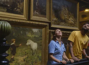 Two people stand on a staircase looking up, with paintings covering the wall behind them