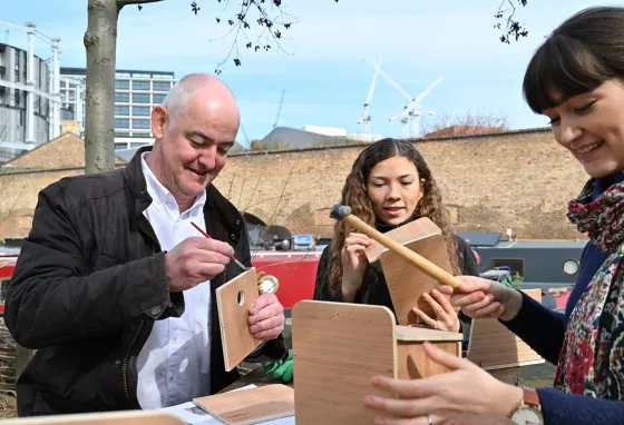 Three people sitting on a picnic table in an urban greenspace, building wooden birdboxes with hammers 