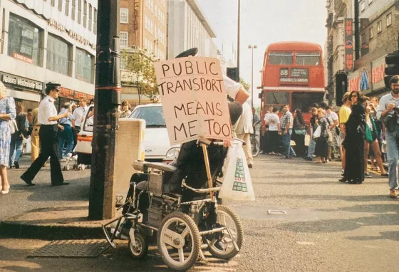 Image of disabled person in 1990s, protesting with plaque saying public transport means me too