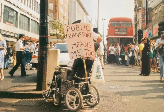 Image of disabled person in 1990s, protesting with plaque saying public transport means me too Image of disabled person in 1990s, protesting with plaque saying public transport means me too