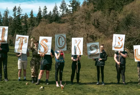 Image of people standing in a line in a countryside setting holding signs reading It's OK 2 B U