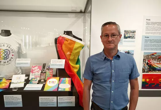 A person standing in front of a glass display of pride flags, t-shirts, leaflets and other memorabilia in a glass case