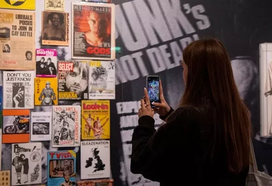 Image of person with long dark hair and dark clothing taking a picture on a mobile of a display of music gig posters at The Museum of Youth Culture