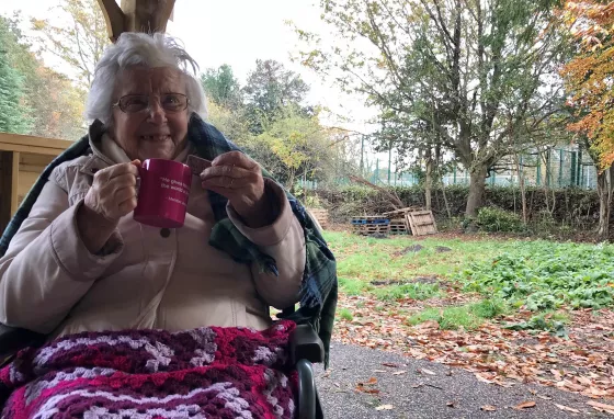 Disability History Month: recognising access in its many forms Elderly woman using wheelchair in garden, smiling with tea and biscuit