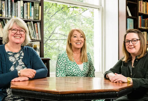 Our Northern Ireland Committee welcomes three new members Three people sit at table in room surrounded by books on shelves. They smile at the camera.