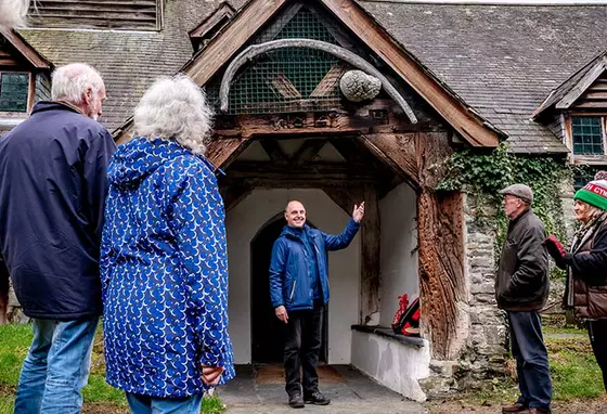 Welcoming visitors to St Tydecho Church in Gwynedd, Wales. Credit: National Churches Trust An outdoor scene of a person welcoming a group of people to a church