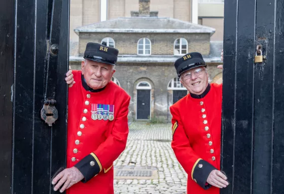 Two people wearing scarlet Chelsea Pensioners' uniform open stable doors at Royal Chelsea Hospital, London with historic building and courtyard behind