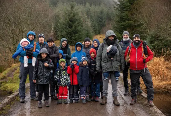 Boots and Beards A large group of people of all ages in walking gear on a rainy day. They are standing on a path through a wooded glen.
