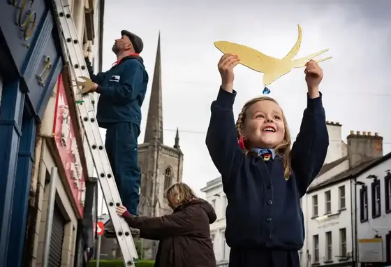 A young girl holds a cut out of a golden bird above her head. In the background are two adults working on a ladder.